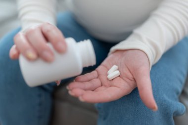 Elinde haplarla plastik kavanoz tutan, hamile bir kadın. Hamilelik boyunca vitamin ve mineral tedavisi görüyor. Evdeki kanepede oturuyor. Yüksek kalite fotoğraf
