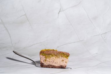 Slice of chocolate Dubai cheesecake with pistachio paste and kataifi covered in chocolate ganache on cake spatula. White marble background with copy space. Side view. Indulgence, gourmet desserts clipart