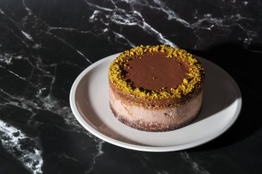 Fıstık ezmeli Dubai çikolatalı cheesecake ve beyaz tabakta çikolata ganache ile kaplanmış kataifi hamur. Kopya alanı olan siyah mermer arkaplan. Hoşgörü, gurme tatlılar, sanat