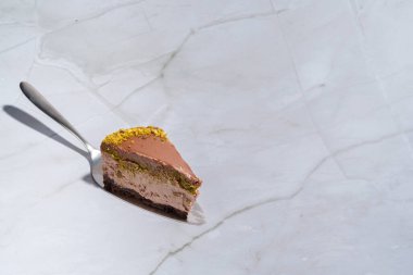 Bir dilim Dubai çikolatalı cheesecake şam fıstığı ezmesi ve kataifi hamuruyla kaplı bir pasta spatulası. Kopyalama alanı olan mermer arkaplan. Hoşgörü, gurme tatlılar, aşçılık.