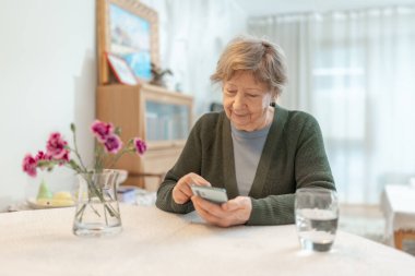Son sınıf öğrencisi bir kadın rahat bir ev ortamında masada otururken akıllı telefon kullanıyor. Teknoloji kavramı, dijital okur yazarlık, çevrimiçi iletişim, bağımsız öğrenme ve aktif yaşlanma. Yüksek