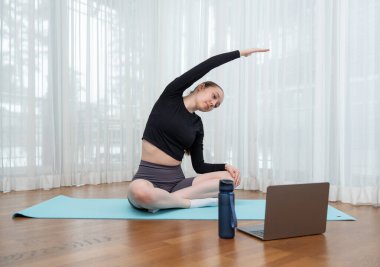 Genç bir kadın dizüstü bilgisayarda egzersiz yaparken yoga minderinde geriniyor, fotokopi çekiyor. Yoga minderinde oturarak esneme egzersizi yapıyorum. Fitness kavramı, esneklik ve sanal eğitim