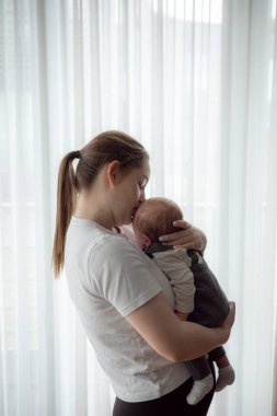Yavru bir bebeği kucağında tutan genç anne evdeki pencerenin yanında duruyor. Erken annelik kavramı, şefkat, bağlılık, ebeveyn sevgisi ve sakin bir ev ortamında bebek bakımı. Yüksek