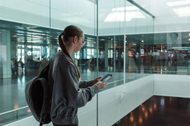 Çağdaş bir havaalanında elinde pasaport ve akıllı telefon tutan sırt çantalı genç bir kadın. Uluslararası seyahat, kimlik doğrulama ve küresel hareketlilik kavramı. Yüksek kalite fotoğraf