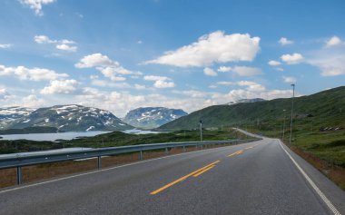 Norveç 'te dolambaçlı bir yol yeşil tepelerin ve kar kaplı dağların arasından geçiyor parlak mavi bir gökyüzü altında tüylü bulutlarla dolu. Bu cennet gibi ortam keşfi davet ediyor.
