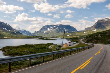 Bereketli yeşillikler, berrak göller ve görkemli dağlarla dolu muhteşem Norveç manzarası boyunca uzanan dolambaçlı bir yol. Parçalı bulutlu gökyüzü huzur ortamına cazibe katıyor.