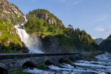 Büyüleyici bir şelale yemyeşil uçurumların üzerine dökülür ve aşağıdaki dolambaçlı yolda bir sis yaratır. Sahne, açık bir günde Norveç 'in doğal manzarasının güzelliğini yakalıyor..