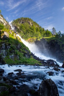 Su, yükselen bir şelalenin üzerinden akar. Güneş ışığı çevredeki yeşillikleri aydınlatırken havada sis yaratır. Rocky arazisi canlı sahneyi çerçeveliyor, doğanın gücünü gösteriyor..