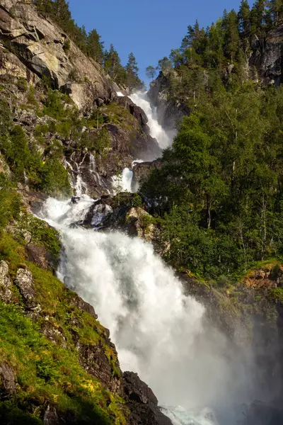 Su, canlı yeşillikler ve yükselen ağaçlarla çevrili kayalık bir arazide enerjik bir şekilde akar. Parlak güneş ışığı bu doğal harikanın güzelliğini arttırır..