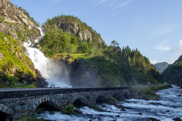Şelale dramatik bir şekilde kaya oluşumları üzerinden aşağıdaki bir nehre akar, canlı ağaçlar ve tepelerle çevrilidir. Bir taş köprü nehri boydan boya geçerek, huzurlu manzarayı güçlendirir..