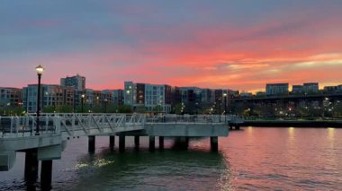 Weehawken, NJ, USA, 19 Nisan 2025: canlı sarı, turuncu ve kırmızı renklerle Weehawken üzerinde gün batımı. Gökyüzü renkleri suya yansıyan iskeleden görünüm.