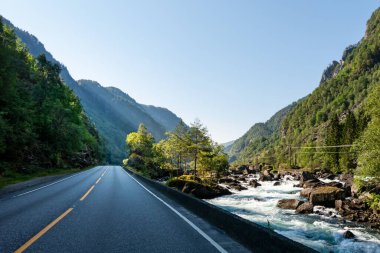 Norveç 'te dolambaçlı bir yol, berrak gökyüzünün altında yemyeşil dağlarla çevrili canlı bir nehrin yanında uzanır. Güneş ışığı doğanın sükunetini vurgulayarak manzarayı aydınlatır..