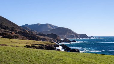 Rocky Point 'ten Kaliforniya' ya uzanan Big Sur 'daki canlı manzara dramatik kıyı şeridi boyunca yeşil çimenler içeriyor. Dalgalar kayalara çarparken uzak dağlar ufku oluşturur..