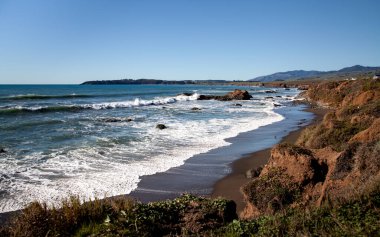 Dalgalar Cambria, California 'da kayalık bir kıyı şeridine çarpar, açık mavi bir gökyüzü ve uzak tepelerle.