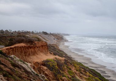 Kaliforniya, Carlsbad 'daki kumlu sahile vuran dalgalar kıyı erozyonunu canlı toprak tonlarını ortaya çıkarıyor. Pasifik Sahil Otoyolu bulutlu bir gökyüzünün altında kıyı şeridine paralel ilerliyor..