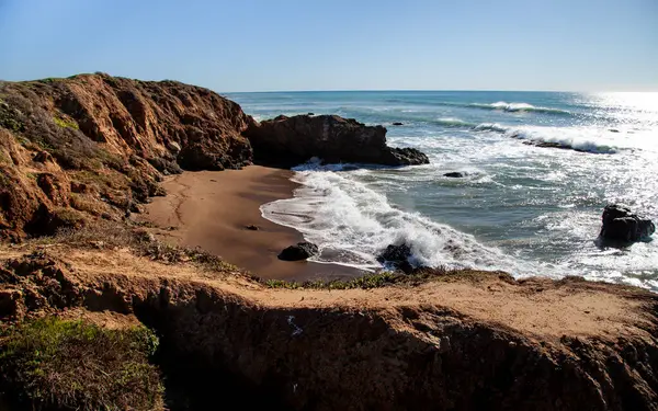 Dalgalar engebeli uçurumlara çarparak güneşli bir günde açık mavi gökyüzünün altında kaya oluşumları arasında uzanan kumlu bir sahili ortaya çıkarır. Huzurlu sahne doğanın güzelliğini yakalar..