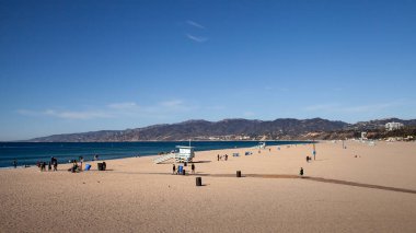 İnsanlar kış boyunca açık mavi bir gökyüzü altında Santa Monica Sahili 'nde rahatlar ve çeşitli aktivitelere katılırlar. Cankurtaran kulesi kumlu sahile ve nazik dalgalara bakıyor..