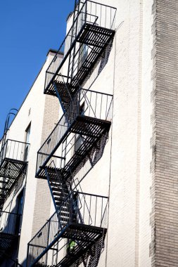 Spiral şeklindeki yangın merdivenleri tuğla bir binaya bağlı. Siyah metal yapısı açık mavi gökyüzüne karşı duruyor, mimari tasarımı vurgulanıyor..