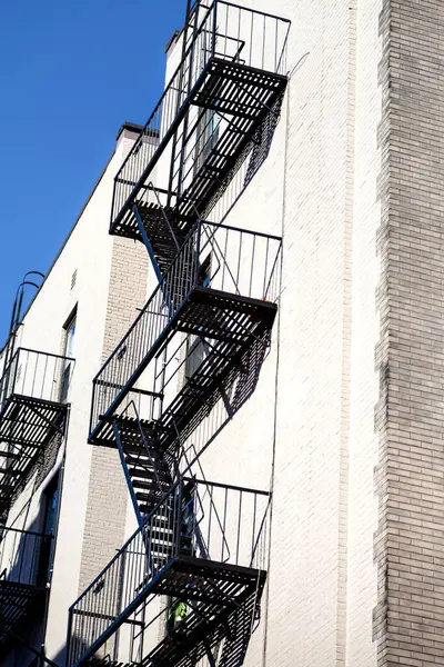 Spiral şeklindeki yangın merdivenleri tuğla bir binaya bağlı. Siyah metal yapısı açık mavi gökyüzüne karşı duruyor, mimari tasarımı vurgulanıyor..