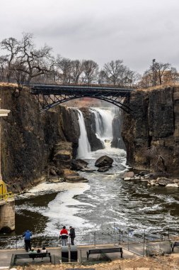 15 Mart 2025, Paterson, New Jersey, ABD: İnsanlar ilkbaharın başlarında Büyük Şelale 'yi gözlemlemek için platformda dururlar. Dramatik uçurumlar ve yaya köprüsü çerçevesi bulutlu sahne.