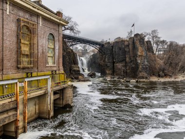 15 Mart 2025, Paterson, New Jersey, ABD: Great Falls şelalesi ve bulutlu bir gökyüzünün altındaki tarihi hidroelektrik santrali geniş görüşlü. Engebeli uçurumlar ve arka planda görünen Amerikan bayrağı. 