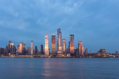 Weehawken, NJ, ABD 19 Nisan 2025: Manhattan silueti gün batımından sonra Hudson Yardları ile birlikte, sakin Hudson Nehri 'nde parlayan kuleleri ve yansımaları aydınlatan ışıklar. 