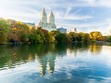 New York 'taki Central Park' ın renkli yeşillikleri ve parlak gökyüzünün altındaki sakin göl suyuna yansıyan ikonik bina kuleleriyle manzarası..