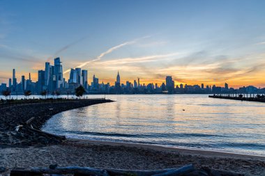 Manhattan silueti üzerinde canlı bir gün doğumu Hudson Nehri boyunca Hoboken, NJ 'den izleniyor, parlayan bulutları yakalıyor ve sabahın erken saatlerinde suya yansıyan ışığı yansıtıyor..