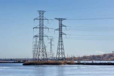 Yüksek elektrik hatları, donmuş suyun üzerine yansıyarak mavi kış gökyüzünde uzanıyor. Manzara, elektrik ağının ölçeğini vurgulayarak, alt kısımda bataklık barındırıyor..