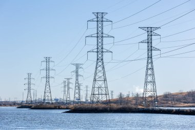 Kablo hatları olan, bataklık ve mavi gökyüzü kontrastlı bir güç iletim kuleleri dizisi. Önde gelen perspektif, enerji altyapılarının ölçeği ve derinliğine dikkat çekiyor.