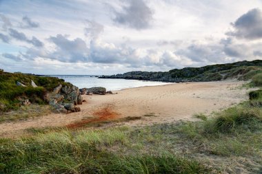 Norveç, Farsund 'da kayalık çıkıntılar ve canlı yeşilliklerle çevrili sakin bir plajın kıyısında hafif dalgalar tur atar. Gökyüzü yumuşak bulutlarla süslenmiş, alacakaranlıkta huzurlu bir ortam yaratıyor..