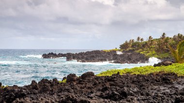 Hana, Maui, Hawaii 'deki Waianapanapas lav kayalıkları boyunca manzaralı bir manzara, Pasifik Okyanusu dalgalarının engebeli kıyı şeridine doğru siyah volkan kayalarıyla çarpışını gösteriyor.. 