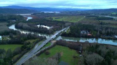 Yüksek hava manzarası, Valdivia, Şili yakınlarındaki kırsal bir bölgede dolambaçlı bir nehirden geçen manzaralı bir köprüyü gösteriyor. Nehir yemyeşil alanlar ve ormanlar arasında kıvrılır, bulutlu gökyüzünü yansıtır.