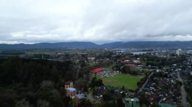 Valdivia şehrinin panoramik hava manzarası, çevresindeki yeşil kırsal alanla sorunsuz bir şekilde harmanlanmış kentsel genişliğini gösteriyor. Görüntü bulutlu bir gökyüzünü yakalıyor, benzersiz