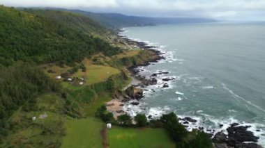 Yeşil tepeler ve Pasifik Okyanusu 'yla buluşan uçurumların olduğu engebeli bir kıyı şeridini gösteren manzaralı bir hava aracı. Kıyı şeridi küçük kır evleri ve çiftliklerle kaplı. Bu da kırsal yaşamın harmanlandığını gösteriyor.