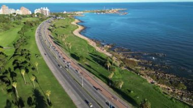 Montevideo manzaralı kıyı yolunun geniş bir hava görüntüsü iyi korunan bir park alanı boyunca uzanıyor. Palmiye ağaçları ve yeşilliklerle kaplı geniş yol kayalık kıyı şeridi boyunca uzanır.