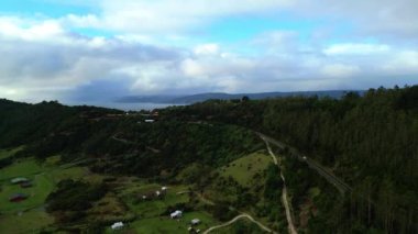 Uzak sahile doğru uzanan yemyeşil bir yamaç manzarası. Virajlı yollar ve dağınık küçük evler uzak kırsal yaşam hissi veriyor. Tepelerin canlı yeşilliği.