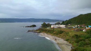 Bir dron Valdivia, Şili yakınlarındaki küçük bir sahil kasabasının güzel bir sahil manzarasını yakalar. Çekimde kayalık çıkıntılar ve yemyeşil tepelerle çevrili kumlu bir sahil sergileniyor. Muhtemelen alçak katlı bir bina.