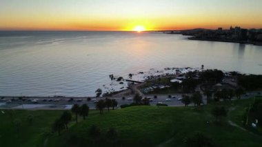 Montevideo Rambla üzerinde gün batımının büyüleyici bir görüntüsü. Önünde yemyeşil bir park ve uzaktan şehrin silueti var. Güneş ufka batıyor, ılık bir portakal döküyor.