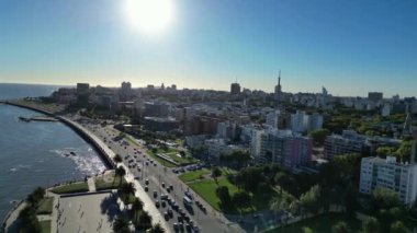Montevideo sahil caddesinin gündüz görüntüsü şehrin sembolik Rambla bölgesinin çarpıcı bir bölümünü yakalıyor. Görüntü kıyı şeridini gösteriyor. Suyun üzerinde güneş yansımaları, modern binalar, yoğun.