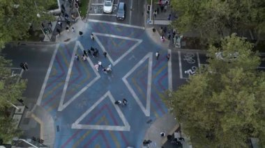 Yaratıcı bir şekilde boyanmış bir kavşağın yukarıdan çekilmiş görüntüsü. Mavi, sarı ve kırmızı geometrik desenler içeriyor. Yayalar kavşakta yürürken kamera yükseliyor.