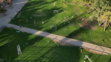 Yeşillik bir alanda çocuklar için bir futbol antrenmanı görüntüsü. Görüntü çoklu küçük çocuk gruplarını gösteriyor. Koçlarının rehberliğinde, dağınık olarak.