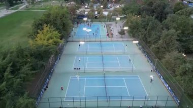 Alacakaranlıkta çekilmiş canlı bir çoklu spor parkının hava perspektifi. Sahnede bir dizi iyi ışıklandırılmış tenis kortları, basketbol kortları ve yemyeşil alanlarla çevrili paten pistleri yer alıyor. Şey...