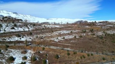 Bu panoramik hava manzarası, parlak mavi bir gökyüzünün altında, benekli bir plato boyunca uzanan dolambaçlı bir yolu gösteriyor. Dağınık ağaçlar ve engebeli arazi huzurlu ve geniş bir manzara yaratır.