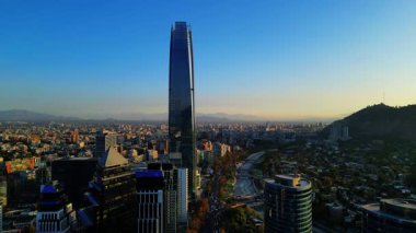Güney Amerika 'nın en yüksek binası olan Gran Torre Santiago gökdeleninin gün batımında şehrin ufuk çizgisinde yükseldiğini gösteren İHA videosu. Görüntüler And Dağları 'nı uzaktan yakalıyor.