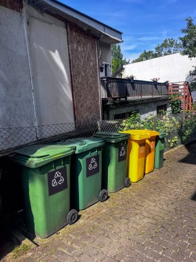Fotoğrafta kaldırım kenarındaki bir sıra çöp konteynırının şehir manzarasının arka planına karşı görüntüsü yer alıyor.