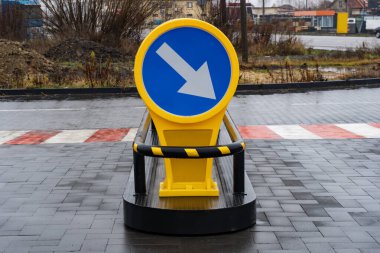 Parlak sarı tabela araçların sağa dönmesi yönünü gösteriyor. Islak ve kısmen gelişmiş bir alana yerleştirilmiş.