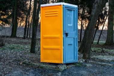 Canlı mavi ve turuncu renkli portatif bir tuvalet gün batımında gün batımına doğru kaybolurken ağaçların arasında yer alır.