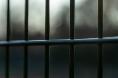 Alacakaranlıkta bulanık bir arkaplan karşısında yeşil metal bir çitin yapısını yakalıyor, çizgileri ve derinliği vurguluyor.