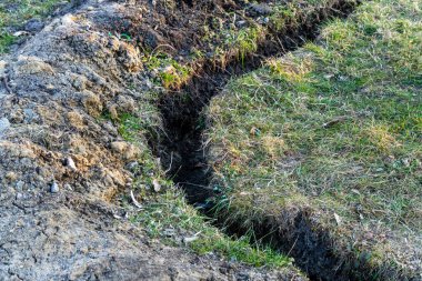 Çimenli bir alanda bir çukur kazılıyor, bu da peyzaj ya da kamu hizmetleri için hazırlık olduğunu gösteriyor. Toprak dönüyor ve çimenler bozuluyor.
